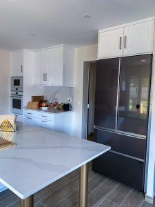 Mobiliário de cozinha branco com armários de estilo clássico, frigorífico americano embutido e bancada em quartzo com veios cinza.