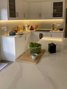 Bancada de cozinha em quartzo branco com veios cinzentos, iluminação LED sob os armários superiores e acabamento lacado.